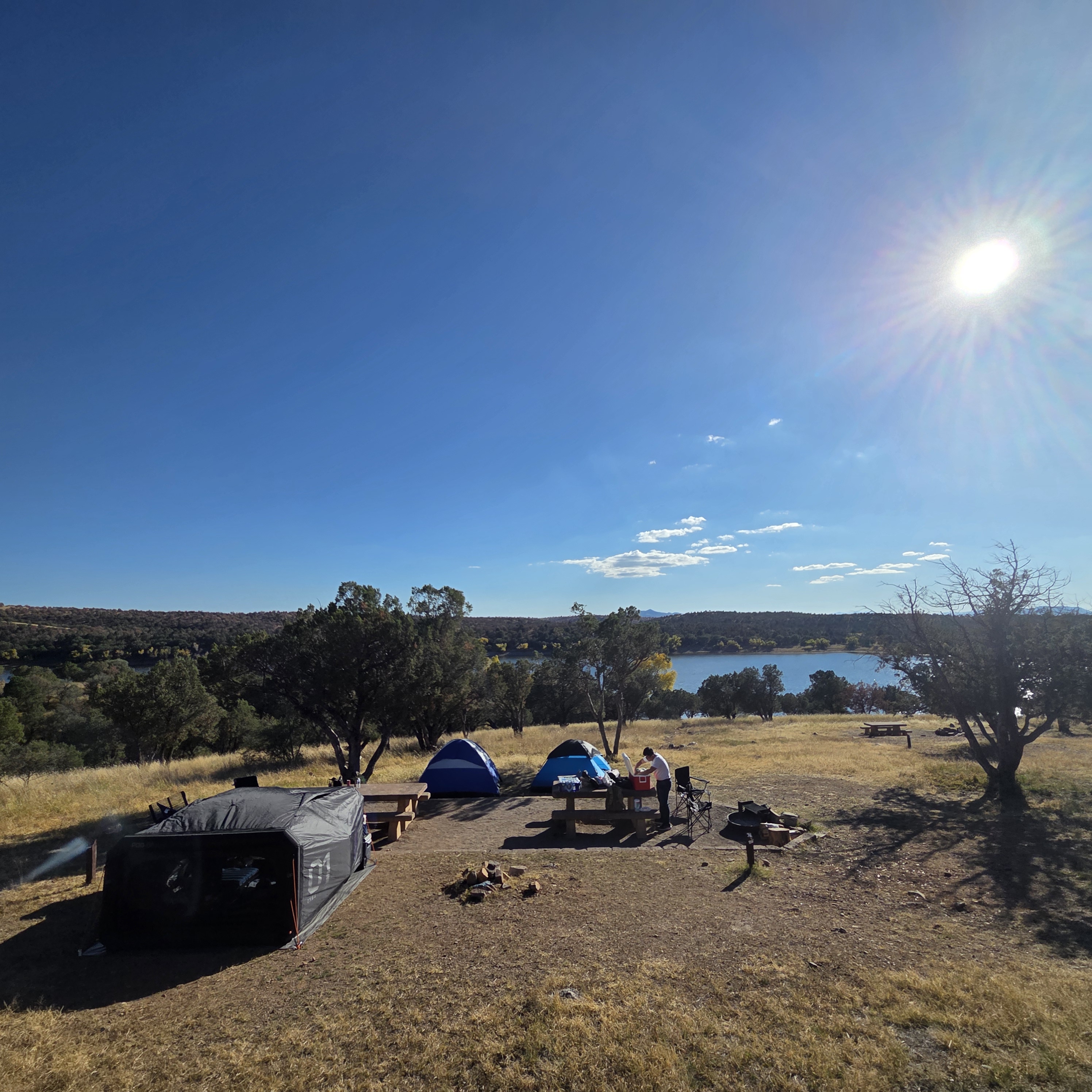 horizon photo of a campsite with a black rectangular infaltable tent and 2 smaller tents across it with the sun high in the sky