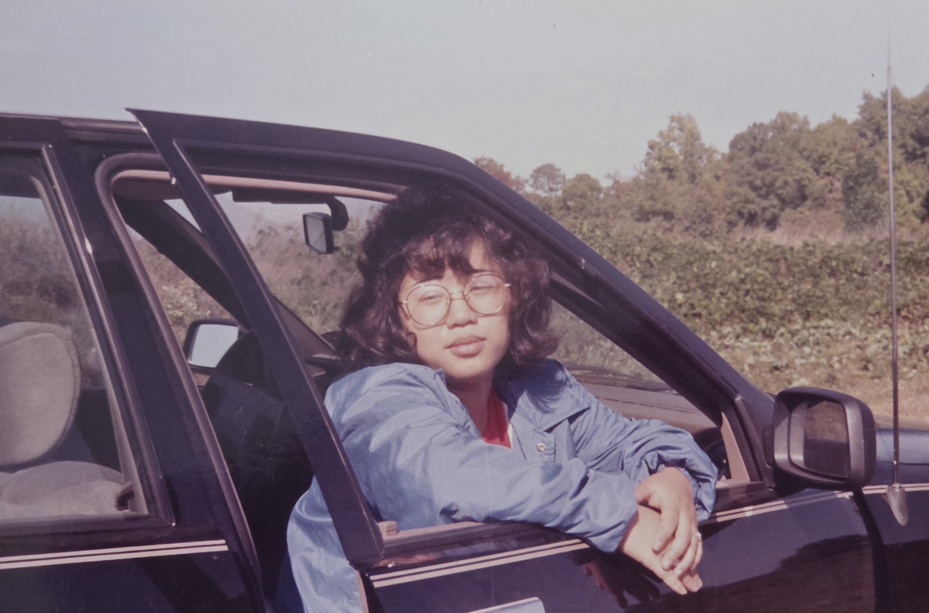 retro photo of a beautiful women sitting with door partially opened and her arms leaning on opened window of car looking in the distance